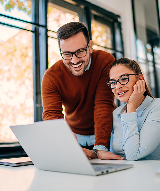 Two people wearing glasses, a man in an orange sweater and a woman in a light blue shirt, smiling as they look at a laptop together in a bright, modern office with large windows.