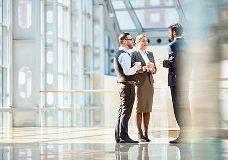 Three business professionals in formal attire stand in a bright, modern office space, engaged in conversation. Sunlight streams through large windows, creating a professional and open atmosphere.