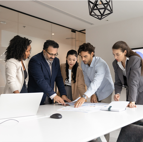 Five business professionals stand around a conference table, collaborating and reviewing documents together in a modern office setting. Laptops and papers are on the table.