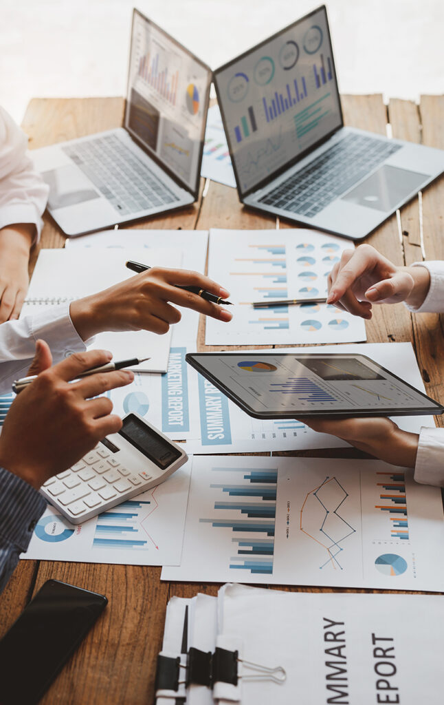 Four people work together at a table covered with charts, graphs, and reports. Laptops display data, and hands point to documents and a tablet, suggesting analysis and discussion of business or financial statistics.