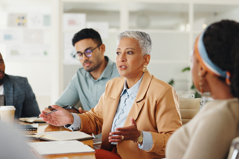 A woman with short gray hair, wearing a tan blazer, speaks in a meeting with three colleagues. She gestures with her hands, and notebooks and coffee cups are on the table in front of them.