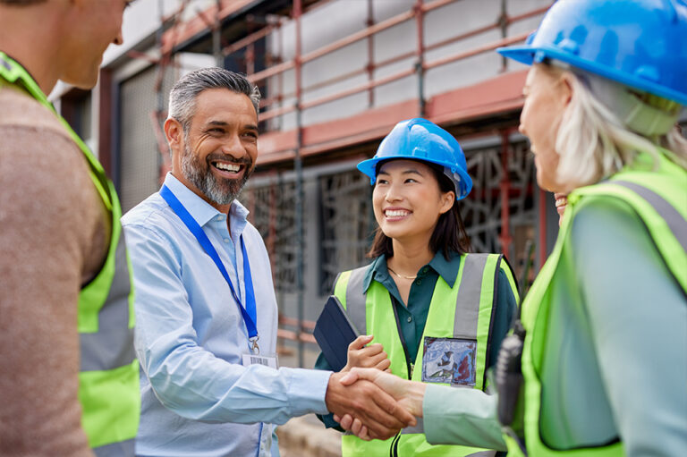 A man in business attire shakes hands with a woman in a hard hat and safety vest at a construction site. Two other people in safety gear stand nearby, all smiling and engaged in conversation.