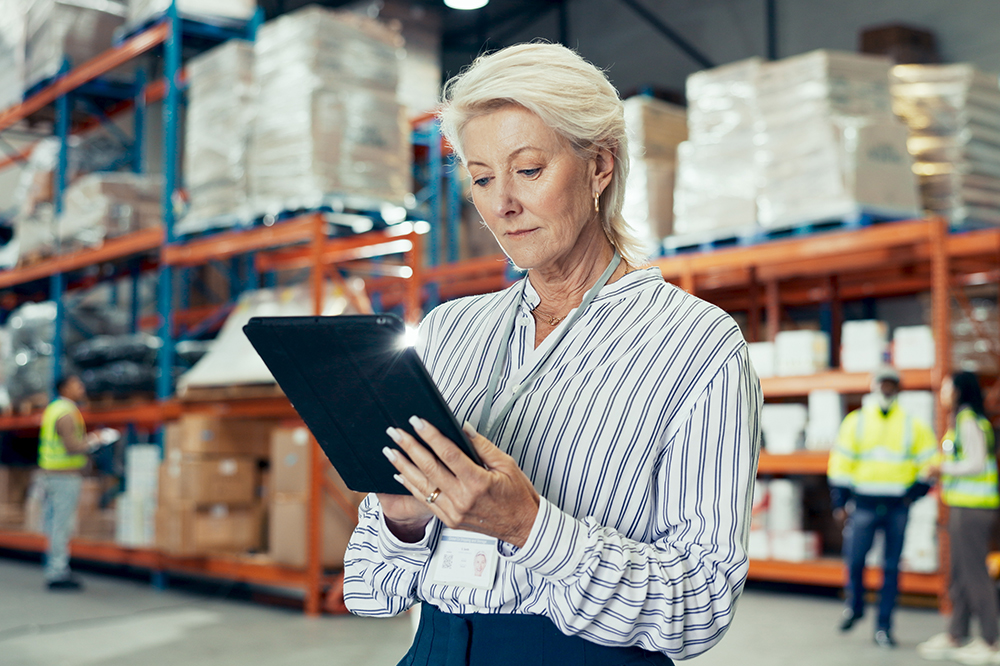 A woman with short blonde hair, wearing a striped shirt and lanyard, uses a tablet in a warehouse with shelves stacked with boxes and two workers in safety vests in the background.