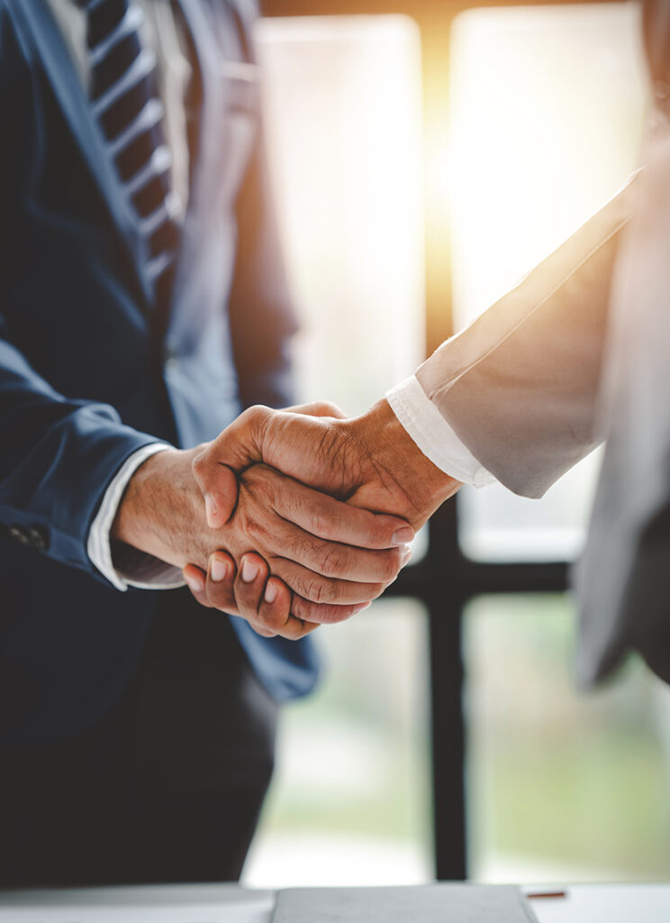 Two people in business suits shaking hands, symbolizing agreement or partnership, with sunlight shining through windows in the background.