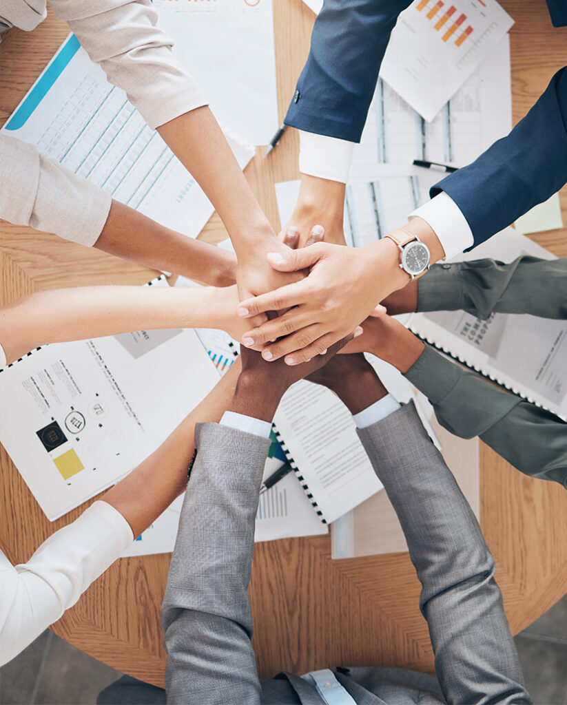 Six businesspeople stack their hands together in the center of a table covered with documents, graphs, and charts, symbolizing teamwork and unity in a professional setting.